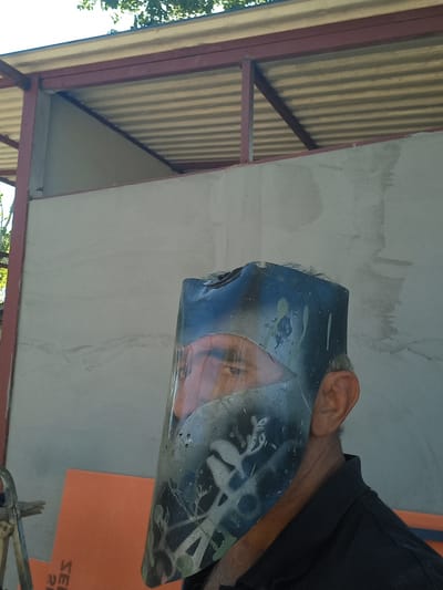 A man wears a welding mask over his face, obscuring most of it. The mask is dark and appears to be spray-painted with white designs. Only his eyes and part of his left ear are clearly visible. He is wearing a dark collared shirt. The background consists of a light-colored wall and a corrugated metal roof supported by red beams. In the lower left corner, a partial view of a metal ladder and an orange object with white lettering are visible. The overall scene suggests an outdoor setting, possibly a construction site or workshop. The presence of overhead trees indicates a natural element to the environment. The location is identified as Çatalca, Turkey.