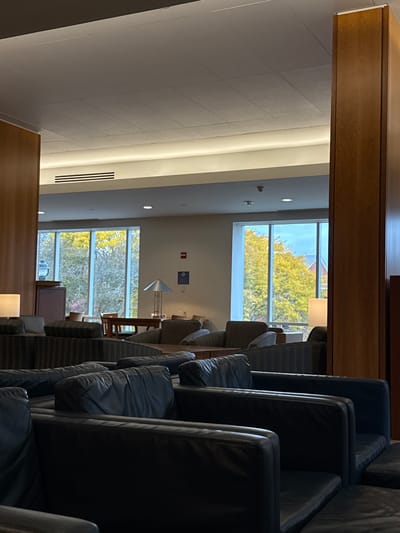 This image depicts the interior of a common area or lounge within the Baker Library at Bentley University in Waltham, Massachusetts, United States. The scene is brightly lit, combining natural light from large windows with internal artificial lighting.
In the immediate foreground, several rows of unoccupied dark, possibly black, leather armchairs are visible, extending towards the midground. Beyond these, various groupings of fabric-upholstered armchairs and sofas, featuring striped patterns (likely dark blue/grey and black), are arranged around dark wooden tables. On the left side, near a window, a dark wooden cabinet or credenza is present, with a table lamp featuring a white rectangular shade. On a table in the mid-right, a modern metallic table lamp with a conical shade stands.
The background is dominated by a series of large, multi-pane windows that offer a view of vibrant yellow autumn foliage on trees, suggesting the season. Through the rightmost window, a portion of a red brick building with white trim is discernible. On the light-colored wall between the windows, a small rectangular red sign (likely a fire extinguisher marker) and a square blue informational sign are affixed.
The ceiling consists of light-colored acoustic tiles, equipped with recessed lighting fixtures, a prominent linear light strip running along the wall, air vents, and smoke detectors. Tall, light-toned wooden columns or partitions frame the scene on both the left and right sides, contributing to the architectural structure. No individuals are present in the image, rendering the space quiet and unoccupied.