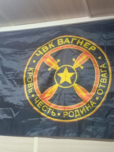 The image shows a dark blue flag with a circular emblem. The emblem is bordered in yellow and red, and features a yellow star in the center, surrounded by four crossed swords and a wheel-like design. Text in yellow Cyrillic letters encircles the emblem. The top portion of the text reads "ЧВК ВАГНЕР" (ChVK Vagner), which translates to "PMC Wagner". The bottom portion of the text reads "ЧЕСТЬ • РОДИНА • ОТВАГА" (Chest' • Rodina • Otvaga), meaning "Honor • Motherland • Courage". The flag is displayed against a plain background, appearing to be indoors. The lighting suggests it is daytime. There are no people or specific scene elements visible beyond the flag itself. The image is a close-up of the flag, with slight wrinkles and folds visible, indicating it is made of fabric.
