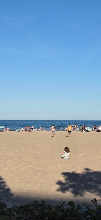 The image depicts a beach in El Port de Sagunt, Spain, with a clear blue sky over the water. The sandy beach is populated with people enjoying the summer weather, indicated by umbrellas and beachgoers. A small child is visible in the foreground, and shadows cast by trees fall on the sand near the right edge of the frame. The image is vertically oriented, with the sky occupying the left half and the beach covering the right half.