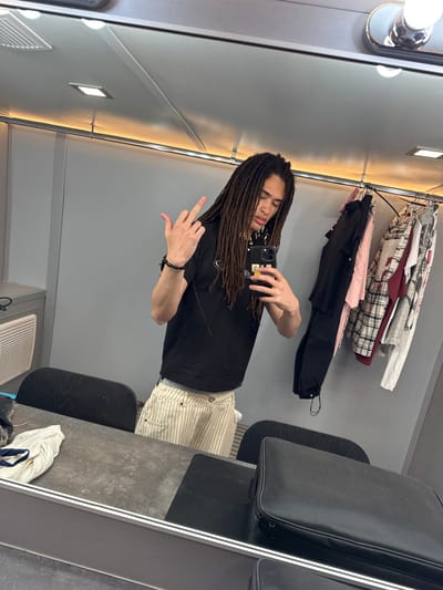 A person with long dreadlocks is taking a selfie in a dressing room. They are wearing a black t-shirt and beige and white striped pants, and holding a smartphone. The person is giving the middle finger. The dressing room contains a clothing rack with various garments hanging on it, a suitcase, and a vanity with a mirror and lights. It is not possible to infer the location of San Bartolomé, Spain from the image content.