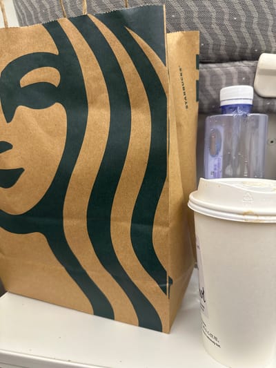 The image displays a close-up of a Starbucks paper bag with its iconic green siren logo. To the right of the bag, a white paper coffee cup with a white lid is visible, showing some coffee stains. Behind the cup, a clear plastic bottle with a blue cap and condensation on its surface is partially visible. The background appears to be a textured grey fabric, possibly upholstery. The scene is set on a white surface, which could be a table or counter. The context of Guangming District, China, is not directly inferable from the visual elements within the image itself.
