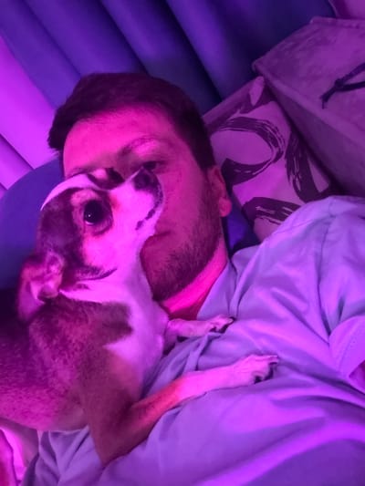 A low-angle, close-up shot captures a person and a dog nestled together, illuminated by pink and purple lighting. The person, appearing to be male with dark hair and stubble, lies on his back with his head tilted upwards. His left arm is bent, and his hand rests on his chest. A small dog, with white and brown markings, is held close to the person's chest, with its head resting on the person's chin. The dog's eyes are open and looking upwards. The background consists of draped purple fabric, likely curtains, and a patterned pillow. The scene suggests an intimate and relaxed moment between the person and the dog. The image does not provide any information about the location in Tokaryovo, Russia.
