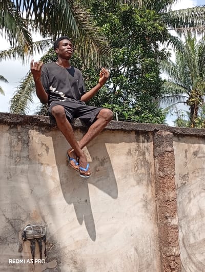 In Osina, Nigeria, a young adult male with dark skin is depicted sitting atop a weathered concrete wall under daylight conditions. He wears a black t-shirt and black shorts, with blue and brown flip-flops on his feet. His legs are crossed at the ankles and dangle over the wall's edge, casting distinct shadows on its surface. His arms are bent and raised, hands open with palms facing upwards. His head is slightly elevated, and his gaze is directed towards the upper right, away from the camera. The background features dense tropical foliage, including prominent palm trees, under a partially visible sky. A damaged, cage-style outdoor light fixture with exposed wiring and dark residue is affixed to the lower left section of the concrete wall. The image bears a 'REDMI A3 PRO' watermark in the bottom left corner.