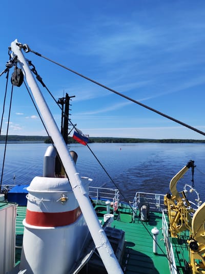 The image presents a bright, sunny daytime scene from the deck of a ship cruising on a wide body of water. The perspective looks forward and slightly to the right, towards a distant, forested shoreline under a clear blue sky.
In the immediate foreground, various parts of the ship are visible. A large white mast or boom with associated pulleys, thick cables, and ropes dominates the left side of the frame, extending diagonally. Below it, a white structure with a distinct red band, possibly a funnel or vent, is prominent on the green-painted deck. To the right, part of a yellow crane or davit is visible. White railings line the edges of the deck. Further back on the ship, a Russian flag, displaying its white, blue, and red horizontal stripes, flies from a mast, indicating the vessel's nationality and the setting in Russia, specifically near Анхимово. A blue box on the deck shows faint white Cyrillic text, possibly "АПК-ННК".
The broad expanse of water in the midground is deep blue, with ripples and a noticeable wake suggesting the ship is actively moving. Two small red buoys are seen floating in the distance. The background is dominated by a flat, green line of densely forested land, indicating a riverbank or lake shore. Above the horizon, the sky is a vibrant blue with only a few very light, wispy clouds.
No people are visible in the photograph. The scene conveys a sense of peaceful travel on a clear, warm day on a significant waterway.