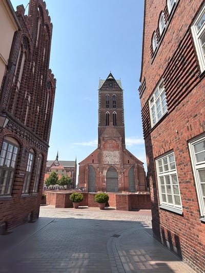 The image shows a view down a narrow street in Wismar, Germany, leading to a prominent brick church with a tall tower. The street is paved with cobblestones and lined with brick buildings on either side. On the left, a tall, narrow brick building with arched windows and decorative brickwork dominates the foreground. On the right, a wider brick building with multiple windows is visible. In the distance, the church, with its Gothic architecture and spire, stands out against a clear blue sky. Planters with round green bushes are placed along a low brick wall in front of the church. The overall scene depicts an urban environment with historical architecture.