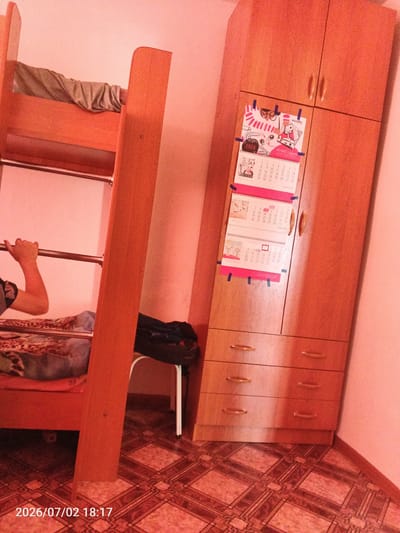 This is a candid, slightly blurry, full-shot photograph taken indoors, likely in a bedroom, as suggested by the presence of a bunk bed and a wardrobe. The scene is bathed in a warm, reddish-orange light, which casts a reddish hue over everything, including the walls and the furniture. The dominant objects in the frame are the wooden bunk bed on the left and a tall wooden wardrobe on the right.
The bunk bed appears to be made of light-colored wood. The top bunk is partially visible with a crumpled gray blanket. A person's arm and hand are seen grasping the metal railing of the bunk bed, suggesting they are either climbing or using it as a support. Below the bunk bed, a patterned blanket is visible on the lower bunk, along with some dark clothing or fabric items.
The wardrobe is a tall, wooden unit with two doors at the top and three drawers at the bottom. On the upper doors of the wardrobe, several calendars are attached with blue tape. These calendars show various illustrations and dates, with some text visible, including "2026", "BOYARD", and what appears to be Russian text. One calendar clearly shows the month of January 2026.
The floor is covered with patterned tiles, predominantly in shades of brown and beige, with a geometric design. A black bag is placed near the bottom of the wardrobe, and part of a white stool is visible next to it.
The time stamp at the bottom of the image indicates "2026/07/02 18:17," suggesting the photograph was taken in the evening of July 2, 2026. The overall atmosphere feels domestic and lived-in. The color cast and the style of the furniture might indicate a location in Russia, consistent with the provided location context.