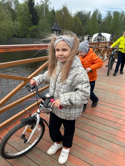 A young girl is standing on a wooden pier, gripping the handlebars of a white bicycle. She wears a plaid jacket, black pants, white sneakers, and a gray headband. Her hair is blonde with some pink streaks. Behind her, another child in an orange jacket lies prone on the pier. Further in the background, a person in a yellow jacket stands near a bicycle. The pier's wooden planks are visible, and a wooden railing separates the pier from the water. Trees are visible across the water, and a white building is partially visible in the distance. The location is Grebnevo, Russia.
