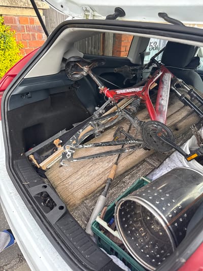 The image displays the open rear cargo area (boot/trunk) of a white car, heavily loaded with various items. The vehicle appears to be a hatchback or small SUV, with "Ford" faintly visible on the trim near the boot hinge.

The primary subjects filling the boot are a red bicycle, partially disassembled, with "BARRACUDA" and "TREMOR" printed on its frame, and a large, perforated stainless steel drum, typical of a washing machine. Beneath these, several rough wooden planks or parts of a pallet are stacked, along with what appears to be another black bicycle frame and other bike components like forks and pedals. A green plastic crate is visible underneath the metal drum, and a long object with a cork-like handle, possibly a fishing rod or tool, is also present among the clutter. The car's interior shows black carpeted flooring and grey plastic trim.

The activity depicted is the loading or unloading of a significant amount of items, likely for transport, possibly to a recycling center or for disposal.

Notable details include the bright, natural lighting suggesting daytime and dry weather conditions. Outside the car, a brick wall and some green foliage are visible. Through the car window, a number "53" is faintly discernible, possibly a house number. The overall scene conveys a sense of disarray and a practical task in progress. The location context provided is Boston, United Kingdom.