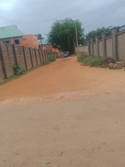 The image depicts a street scene in Gindi Nassarawa Kyarmi Galbi, Nigeria. A paved road is in the foreground, leading into a dirt road lined by a concrete block wall on the left and a decorative concrete wall on the right. The dirt road curves to the left and is bordered by scattered vegetation and debris. On the left side of the dirt road, there are buildings with orange facades and green roofs, one of which has water tanks on its roof. A white van is parked on the dirt road further ahead, near a cluster of trees. The sky is overcast.