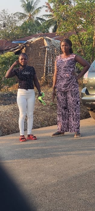 Two adult females are positioned on an asphalt road in Alayi, Nigeria. The female on the left wears a black speckled short-sleeved top, white form-fitting pants, and red open-toed sandals. She holds a pair of bright green footwear or gloves in her left hand, and her right hand is raised near her head. The female on the right wears a pink and black patterned short-sleeved top and matching wide-leg pants with dark open-toed sandals. Her left hand rests on her hip. Both individuals face forward towards the viewer. In the mid-ground, a structure composed of light brown mud bricks with a reddish-brown corrugated metal roof is visible. The background consists of dense green foliage, including palm trees. A dark-colored vehicle, possibly an SUV, is partially visible on the far right, showing its rear fender and tire. The ground beside the road is dry, light brown earth. The scene is illuminated by natural daylight.