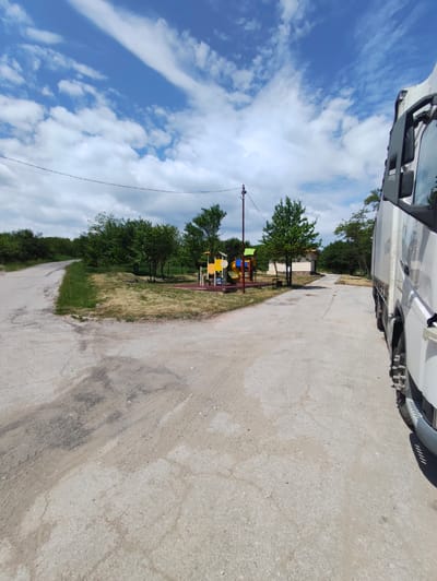 The image presents an outdoor scene at what appears to be an intersection or branching of paved roads in Kostadinkino, Bulgaria. A white truck is parked on the right edge of the frame, partially obscuring the view. To the left and center, a playground with colorful play structures is visible, indicating a recreational area. Beyond the playground, there are trees, vegetation, and a small building. Overhead, a partly cloudy sky dominates the scene. The asphalt surface is cracked and worn, showing signs of use and aging. A power line runs across the sky in the middle of the image.