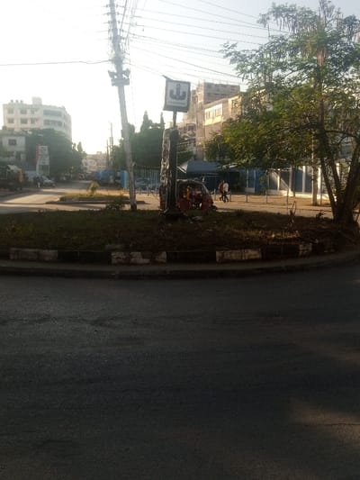 An outdoor, sunlit street scene depicts a roundabout intersection in Shimanzi ward, Mombasa, Kenya. The foreground features a dark asphalt road surface. A raised circular island, serving as a roundabout, occupies the mid-ground, bordered by a black and white painted curb and covered with sparse dry grass and small plants. Positioned on the far side of the roundabout is a leaning utility pole heavily laden with numerous electrical wires. Adjacent to the pole stands a signpost with a white rectangular sign displaying a black anchor symbol and the text "AFRICAN MARINE." A red three-wheeled auto-rickshaw is parked near the base of this signpost.
To the right of the roundabout, two adult males are observed walking away from the camera on a paved area, one wearing a white shirt and the other a darker shirt. Behind them, a blue security fence or barrier is visible, along with mature trees displaying green foliage. In the background, a street extends with multi-story urban buildings of varying architecture on both sides. Additional indistinct vehicles, including a large truck, and several pedestrians are visible further down the street on the left. The sky is bright and partially hazy.