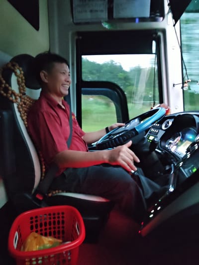A man is driving a bus. He is wearing a red shirt and black pants and has a seatbelt fastened across his chest. He is smiling and looking towards the right. The steering wheel is blue and black and he is holding it with his hands. There is a red plastic basket filled with plastic bags on the floor to his left. The bus is moving as indicated by the motion blur in the background. The interior of the bus is visible, including the dashboard with various lights and displays. The exterior view shows greenery outside the bus, suggesting a rural or suburban location. The image appears to have been taken from the passenger's seat, looking towards the driver. It is possible the location is Pờ Y, Vietnam, but this cannot be confirmed from the image alone.
