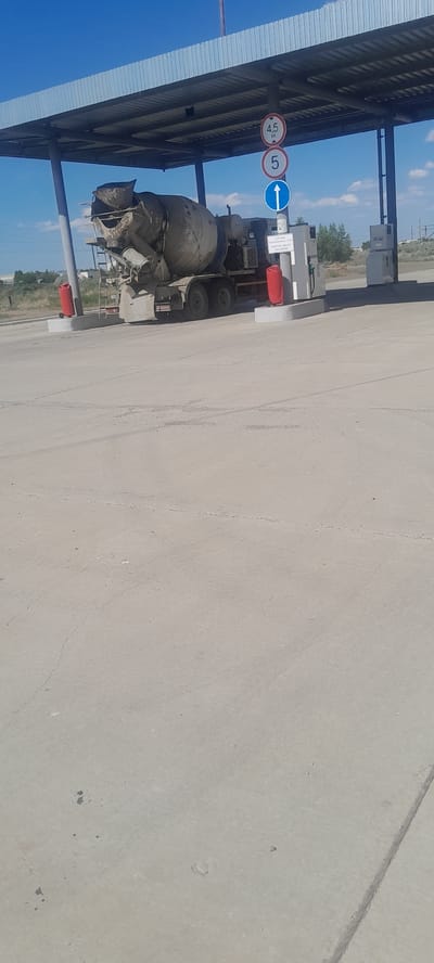 A concrete mixer truck is parked under a covered structure at a fueling station in Kurchatov, Kazakhstan. The truck is gray and appears to be old and dirty. The fueling station has a gray concrete floor and a metal roof supported by white pillars. There are several signs visible, including a sign with "4.5 M" and a speed limit sign with "5". There are also two red posts with cones on top. The sky is blue with some white clouds. The area appears to be rural or industrial.