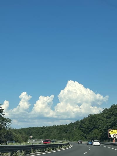 The image shows a bright, sunny day with a clear blue sky on the left and a highway curving to the right. Large, fluffy white clouds are visible against the blue sky. The highway is lined with dense green trees on the right, creating a backdrop of lush vegetation. Several cars are driving on the highway, including a white car, a dark blue car, and a red car. A yellow sign with text is visible above the trees. The scene appears to be on a highway, possibly in a rural or suburban area, given the abundance of trees. The weather is clear and sunny, indicating it is daytime. The angle of the image is slightly tilted. The text on the sign appears to read "FAR petroleum". The image captures a typical highway scene on a pleasant day.