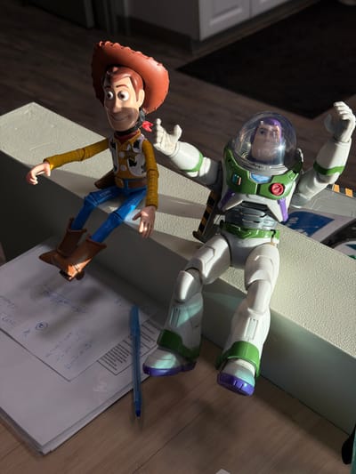 The image shows two Toy Story action figures, Woody and Buzz Lightyear, lying on a stack of papers with a blue pen on top. The paper contains handwritten notes and addresses, including "600 Cessna Dr," "1300 Cornette May," and "3111 Grant McCaracte" which are all located in Delta, British Columbia, Canada. The figures are arranged in a way that suggests they are interacting. They are on what appears to be a light brown wooden surface, with shadows in the background.
