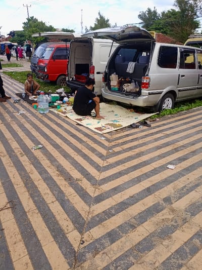 The scene is a paved area in Pameungpeuk, Indonesia, at coordinates -7.668, 107.694. Two men are sitting on a striped mat, one appears to be eating. One man is wearing a black shirt and the other is wearing a blue shirt. There is a silver minivan in the background with its back hatch open. The van is packed with items, including a brown paper bag. The man wearing the black shirt is sitting directly in front of the van. The men appear to be eating and enjoying their time together. There is a large white van and a red minivan parked behind the silver van. There are some other people in the background but they are unclear.  The pavement is striped in black and tan. There are some items on the ground, including a water bottle and a bag.