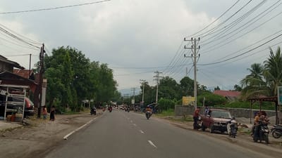 The image depicts a street scene in Beunyot, Indonesia. The sky is overcast with a pale gray, cloudy atmosphere. On the left side of the road, there are buildings with corrugated metal roofs, some appearing weathered. A white structure with an awning is visible, with what appears to be a vehicle and some containers next to it. Two children are standing on the unpaved shoulder of the road.
The main subject is a paved road with white dashed lines marking the lanes, indicating it is a two-way street. The road is populated with several motorcycles, some carrying one or two riders, traveling in both directions. A red pickup truck is parked on the right side of the road, facing away from the viewer. Several people are standing near the truck. Further down the road, more motorcycles and a few cars are visible.
Electrical poles with numerous wires crisscross the scene, a common feature in the urban landscape. Lush green trees and foliage line the sides of the road, interspersed with houses and some commercial structures. A billboard with yellow text is mounted on a concrete wall on the right. The overall impression is of a busy, lived-in street in a tropical or subtropical region.