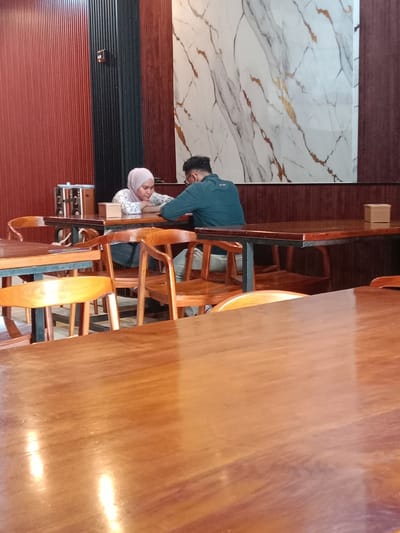 The image depicts two people seated at a table in a restaurant. A woman wearing a light pink hijab and a patterned blouse is sitting across from a man with dark hair and glasses, wearing a blue shirt. They are both leaning on the table. The restaurant has wooden tables and chairs. The background includes a red wall with a black ribbed section, and a marble-patterned decorative panel. The location is Pandean, Indonesia.