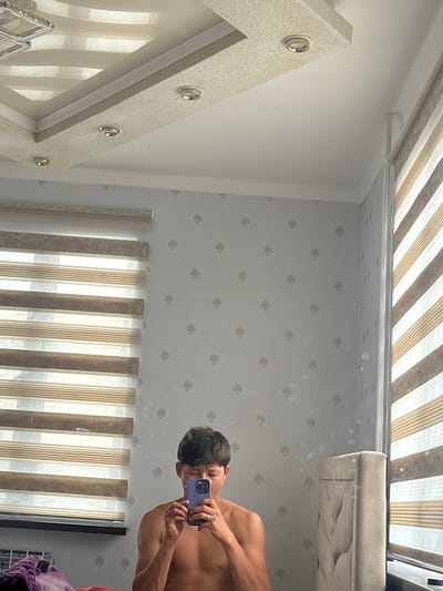 The image depicts an interior scene, likely a bedroom, with a young man taking a mirror selfie. He is shirtless and holding a purple smartphone in his right hand, with his left hand resting on his chin. The room features a textured ceiling with recessed lighting fixtures and a decorative molding. The walls are covered in light gray wallpaper with a subtle floral pattern. To the left, a window is covered by striped blinds. To the right, a portion of a tufted headboard is visible. The overall impression is of a domestic setting.