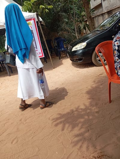 The image depicts an outdoor scene in Okpatu-Ukehe, Nigeria. A person, viewed from behind, stands on a sandy, light brown earthen surface. They are dressed in a long-sleeved white top, a long white skirt, and a bright blue head covering that drapes over their head and shoulders. Dark sandals are worn on their feet. In their right hand, they hold a clear plastic bottle of water with a blue label. A dark shoulder bag is visible on their left side.

To the left, a white canopy structure with support poles is present, partially obscuring a red and white banner displaying partial text, including "O Y" and "UB." Several dark blue plastic chairs are arranged near the canopy. To the right of the central figure, a black sedan car is parked, with its front-left quarter, headlight, and bumper visible. Behind the vehicle, dense green foliage and tree trunks are present, with a light-colored building wall topped by barbed wire visible in the background. In the immediate foreground on the right, the arm and patterned clothing of another individual are partially visible, seated in an orange plastic chair. The scene is brightly illuminated by direct sunlight, casting distinct shadows of figures and overhead vegetation onto the uneven sandy ground, which exhibits numerous footprints and tire impressions.