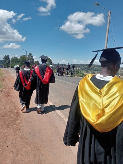 The image depicts an outdoor scene in Waseges ward, Kenya, on a clear, sunny day. The primary subjects are several individuals dressed in academic graduation regalia. In the immediate foreground, facing away from the viewer, stands one person wearing a black gown, a bright yellow satin hood, and a black mortarboard with a tassel. To the left, in the mid-ground, two more individuals also in black gowns and black mortarboards, but with red satin hoods, walk along a reddish-brown dirt shoulder adjacent to a paved road. The individual on the left has visible light-colored pants and athletic shoes, while the one on the right shows light-colored pants and brown shoes.
Further down the asphalt road, which features a solid white line on its edge, a group of approximately 5-7 additional people are visible. Some are standing, and at least two are on bicycles, all appearing to be in similar black graduation attire. On the far left of the mid-ground, a white vehicle is partially visible on the road. The road curves gently to the left and appears to descend into a distant valley.
The environment includes lush green trees and vegetation lining the left side of the road and extending into the background landscape. The sky is clear blue with scattered white cumulus clouds. On the right side of the road, a tall utility pole with a street light fixture extends towards the sky, with visible power lines. Shadows cast by the individuals indicate bright sunlight.