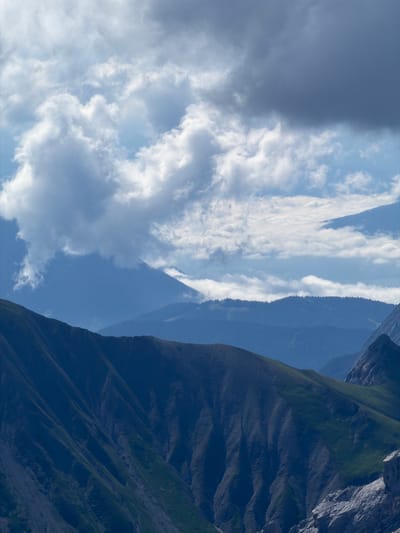 The image displays a mountainous landscape with multiple layers of peaks fading into the distance. The foreground features a steep, dark blue mountainside with visible contours and patches of green vegetation. The middle ground consists of rolling, forested mountains in varying shades of blue. The background reveals further mountain ranges, also in muted blue tones, stretching towards the horizon. The sky above is partially filled with dramatic, volumetric clouds. The upper portion of the image is dominated by a large, dark grey cloud formation, while lighter, white clouds are scattered across the rest of the sky. The scene is natural and appears to be a wide-angle shot of an alpine environment. It is difficult to pinpoint an exact location within Wildermieming, Austria, based solely on the image, but it is clearly a mountainous region typical of the Austrian Alps. There are no people, objects, or actions depicted in the image, only the natural environment.