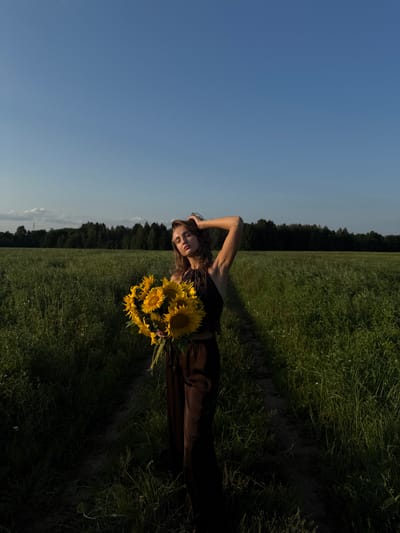 In Aksaitovo, Russia, a young woman stands in a tall grass field under a clear blue sky. She is holding a large bouquet of sunflowers. The woman is wearing a dark brown halter top and matching wide-leg pants. Her eyes are closed, and her right arm is raised, with her hand resting on her head. A dirt path runs through the field behind her, leading towards a dark line of trees in the distance. The lighting suggests it is late afternoon or early evening, with the sun casting shadows across the field.