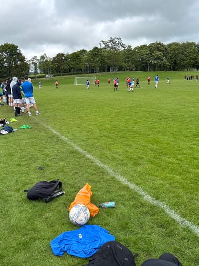A soccer game unfolds on a green field under a cloudy sky. Players in blue and red jerseys are scattered across the pitch. Near the sideline, an orange bag, a soccer ball, a water bottle, a blue shirt, and a black backpack are placed on the grass. A group of individuals stands near the bottom of the frame, observing the game. Trees border the field's edges.