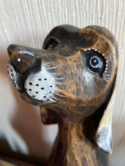 This is a close-up, indoor photograph of a carved wooden figurine of a rabbit. The figurine is primarily brown with carved wood grain visible. The rabbit's face is detailed, featuring a large, dark eye with a white pupil. White paint is used for decorative elements, including dots around the eye, stylized eyelashes, and a cluster of dots on the muzzle, representing whiskers. The nose area is painted white with small black dots. Some white paint is also present on the rabbit's ear. The lighting appears to be artificial, creating a soft glow on the figurine and a slightly blurred background. The background consists of a textured surface, possibly a tablecloth or fabric, with subtle horizontal lines and small, sparkling highlights, suggesting a decorative setting. The overall impression is of a handcrafted decorative item. No people are visible, and no text is present in the image. The context of Габишево, Russia, does not provide any specific visual cues within the image itself.