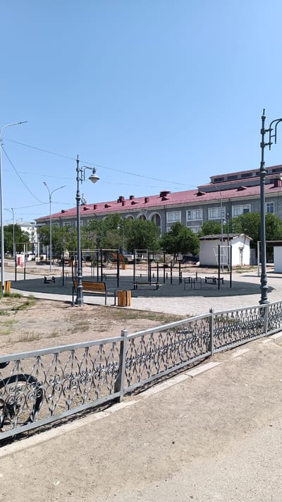 The image depicts an outdoor public space in the city of Balqaş, Kazakhstan, likely a park or recreational area. A long, gray building with a red tiled roof and multiple windows is visible in the background, possibly a school or administrative building. In the foreground, there is a workout area with various pull-up bars and other exercise equipment, set on a dark rubberized surface. Several wooden benches and trash receptacles are also present. A decorative metal fence runs along the edge of the paved walkway in front of the workout area. The sky is clear and blue, and the scene appears to be illuminated by bright sunlight, suggesting daytime. There are no people visible in the image.