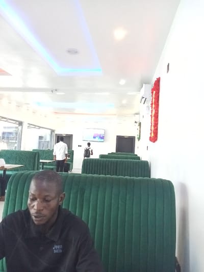 The image shows the interior of a restaurant in Ujevwu, Nigeria. The scene is a dining area, featuring a row of green velvet booths, each with a built-in table. A single man sits facing the camera on the right side of the image, his back to the booth. He is dressed in a black shirt with the inscription "Jean Sale" visible on his right shoulder. Two other men can be seen in the distance, walking towards the back of the restaurant. One wears a white shirt, the other a dark shirt and a black backpack. The restaurant has a white ceiling with recessed lighting, including a long, thin blue light fixture running the length of the ceiling. The walls are white, with an air conditioner visible on the wall to the right of the image. A flat-screen television is mounted to the wall in the back, directly in front of the seated man. The restaurant has a modern aesthetic, with clean lines and a minimalist design.