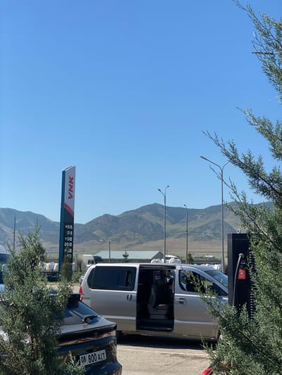This is a medium shot, eye-level, outdoor photograph taken on a clear day. In the foreground, a dark-colored vehicle with a license plate reading "08 800 AIT" is parked. To its right, a silver minivan with its passenger-side door open is parked next to a black charging station. The minivan appears to have its interior lights on. Behind the vehicles, a tall blue and red sign displaying the word "VNK" and gas prices is visible. In the background, a range of green, treed mountains under a clear blue sky. The environment suggests a rural or semi-rural setting, possibly a gas station or charging facility.