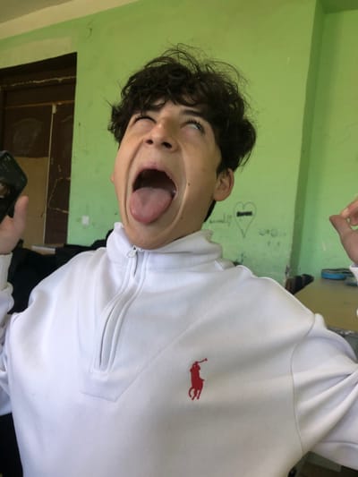 A young male is depicted indoors, reportedly in Ortabatumi, Georgia, performing an exaggerated facial expression. His dark, curly hair is disheveled, and his eyes are rolled upwards, exposing the whites. His mouth is wide open, with the tongue extended. He is wearing a white quarter-zip sweatshirt featuring a red Polo Ralph Lauren logo embroidered on the left chest. A dark-colored smartphone is partially visible in his left hand, while his right hand is partially raised. The background consists predominantly of a light green-painted wall. To the left, a dark brown wooden doorframe and an adjacent dark panel are visible. To the right, faint graffiti, including a heart shape with text, is present on the green wall, with a light-colored surface partially visible at the lower right edge of the frame.