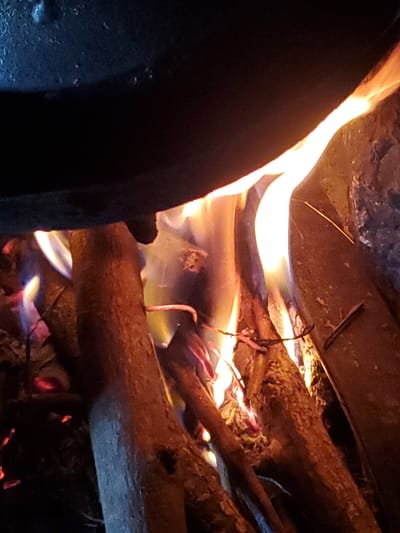 The image depicts a close-up view of a fire burning in what appears to be an outdoor setting. Several pieces of firewood are arranged, and flames are visible, licking around the wood and casting a warm glow. A dark, curved object, possibly a pot or cauldron, is visible in the upper left corner, partially obscuring the flames. The fire is bright and dynamic, with hues of yellow, orange, and hints of blue. The background is dark and indistinct, suggesting it is nighttime or the scene is poorly lit beyond the fire. The location in Kabwir, Nigeria, is not visually evident from the image itself, as there are no discernible landmarks or environmental cues to indicate this specific place. The scene focuses solely on the fire and the surrounding fuel.