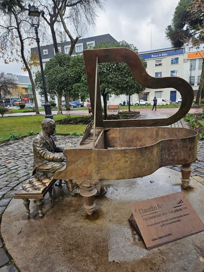 An outdoor bronze sculpture of a grand piano with a seated figure of a man playing it is depicted. The sculpture is located on a cobblestone surface within a park setting, with trees and buildings visible in the background. A bronze plaque is placed on the wet ground in front of the sculpture. The plaque reads "Claudio Arrau" and contains dates and inauguration details in Spanish, indicating the location is Chillán, Chile, with the inauguration date of February 2024. The man in the sculpture is wearing a suit and is seated on a bench in front of the piano. The piano is open, revealing its interior. The environment appears to be a public space, possibly a square or plaza, with overcast skies.