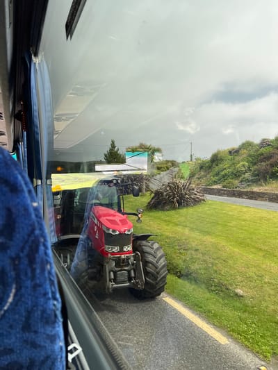The image, taken from inside a vehicle, displays a red Massey Ferguson tractor parked on the side of a road in Dunfanaghy, Ireland. The tractor is the central focus, positioned parallel to the road with its front facing towards the viewer. A yellow line is visible on the asphalt road, separating the lane from the grassy verge. The grassy area is lush and green, with scattered white flowers. Beyond the grass, a stone wall runs along the edge of a property, and a gravel path leads upwards. The landscape is characterized by rolling hills covered in dense, green vegetation, with some scattered trees and bushes. The sky is overcast with diffused light. Reflecting on the vehicle's window, parts of the interior, including a blue seat, are visible.