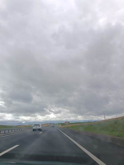 The media file captures a view from a vehicle traveling on a highway in Belarus, likely during daylight hours. The sky is heavily overcast with dark, gray clouds, suggesting a gloomy or potentially rainy day. The road itself is asphalt with clear white lane markings.
The primary subjects are the highway and the surrounding landscape. On the road, a white SUV is prominently visible in the middle lane, with another vehicle further ahead in the distance. To the left of the highway, a metal guardrail separates the road from a grassy embankment, with some sparse vegetation. To the right, a sloping grassy hill rises, dotted with some trees and what appear to be industrial structures or tall chimneys in the distance, particularly a distinct red and white striped tower. A billboard is also visible on the left side of the road.
The overall scene conveys a sense of travel through a rural or semi-rural area. The weather conditions and the vast, empty highway create a somewhat somber or neutral mood. There is no visible text in the image. The activity is straightforward: driving on a highway.