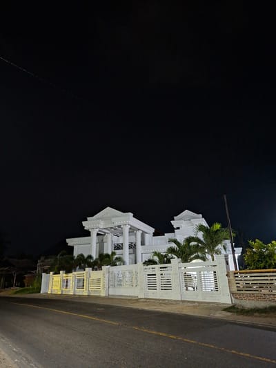 At night in Lancang Barat, Indonesia, a brightly illuminated white building stands adjacent to a paved road with a yellow center line. The building has multiple levels, with decorative lattice work and pillars. Palm trees grow near the building. A power line is visible against the dark sky.