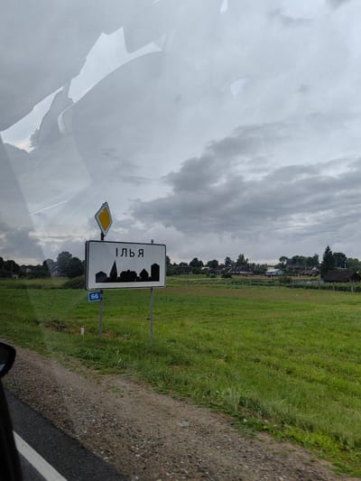 The image depicts a roadside scene under a cloudy sky, likely in a rural area. A signpost with a white rectangular sign bearing the Cyrillic word "ІЛЬЯ" (ILYA) in black letters is prominently featured. Below the text, a black silhouette of houses and a church is visible. A diamond-shaped yellow warning sign is positioned above the town sign. A blue rectangular sign with the number "66" is attached to the signpost at a lower position. The immediate foreground shows a gravel road on the left, bordered by green grass. In the background, a lush green field stretches out, dotted with scattered trees and houses, suggesting a small village or town. The overall atmosphere is subdued due to the overcast weather. The image is captured from a moving vehicle, as evidenced by the reflection on the left side of the frame, which appears to be the interior of a car.