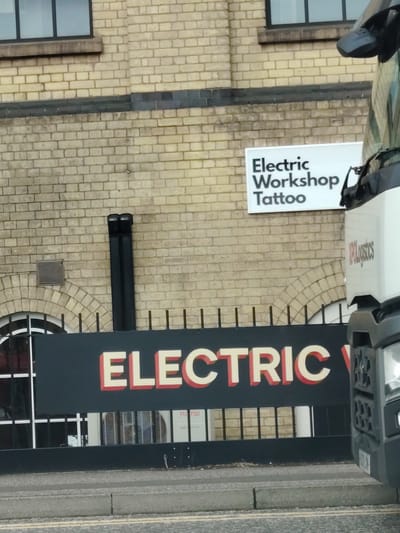 This image captures the exterior of a light-colored brick building, identified as the location of "Electric Workshop Tattoo" on Vicarage Farm Road in Peterborough, United Kingdom.
The upper portion of the building facade features light yellowish-beige bricks with visible mortar lines. Two dark-framed rectangular windows are present in the upper left. Below these, a horizontal band of darker, possibly greyish-blue, bricks extends across the wall. To the right of the windows, a white rectangular sign reads "Electric Workshop Tattoo" in black sans-serif lettering.
On the lower section of the building, an arched window with a dark frame is partially visible on the left, behind black vertical metal railings. Above and to the right of this window, a rectangular metal vent is set into the brickwork. Two black, vertically oriented pipes ascend the wall to the right of the vent.
A large, dark-colored horizontal sign spans the lower part of the building. The word "ELECTRIC" is displayed prominently in large, red, block letters with a lighter outline. The remainder of the sign's text is obscured, but beneath it, a white outdoor air conditioning unit with the "FUJITSU" logo is visible.
The front section of a large white truck is positioned on the right side of the frame, partially obscuring the building and the lower sign. The truck's cab features red "KPLogistics" branding. The dark grey front grille and headlight cluster of the truck are visible in the lower right.
In the foreground, a grey concrete curb separates an asphalt road, which displays faint double yellow lines, from the building's immediate vicinity.