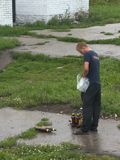 A young man wearing a dark blue t-shirt with the word "ПОЛИЦИИ" (Police) on the back and black pants stands on a wet paved area next to a patch of grass. He is holding a translucent plastic bag in his right hand and appears to be looking down. To his left, on the pavement, are several brown glass bottles with yellow and blue labels, some standing upright and others lying on their sides. Two similar bottles are also visible further left on the pavement. The surrounding environment consists of sparse grass, some wildflowers, and a weathered brick building in the background. The scene suggests an outdoor setting in Miloslavskoye, Russia, possibly a public or semi-public space.