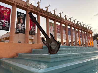 Captured in Los Cristianos, Spain, the image shows the Hard Rock Cafe situated in front of the Palacio de Congresos. A large, metal guitar-shaped Hard Rock Cafe sign is placed in a multi-tiered, light-blue tiled fountain, with water gently cascading down each level. In the background, the Palacio de Congresos, constructed in an ornate architectural style, is visible. The building features a series of vertical columns, topped with statues of archers. Banners with different images are hanging on the columns, likely for promotional purposes. The sky is overcast with a soft light, suggesting it may be either early morning or late afternoon. There are no people visible in the immediate foreground, but there are some in the distance, within the banners on the building.