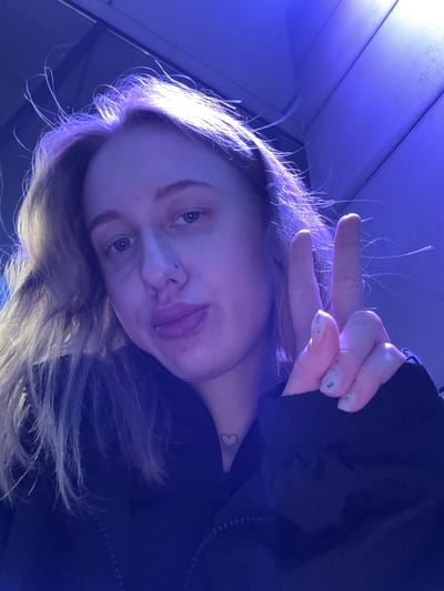 A selfie of a young white-skinned person with blonde hair. The person is holding up a hand, making a peace sign, with light blue nail polish. They have blue eyes, a nose piercing on the left side, and are sticking their tongue out slightly. A small heart tattoo is visible on their neck. The background appears to be indoors, possibly a room with blue lighting and white walls. It is impossible to verify a specific city, such as Ol'ginskaya, Russia, or any location without additional context or visual cues in the image.