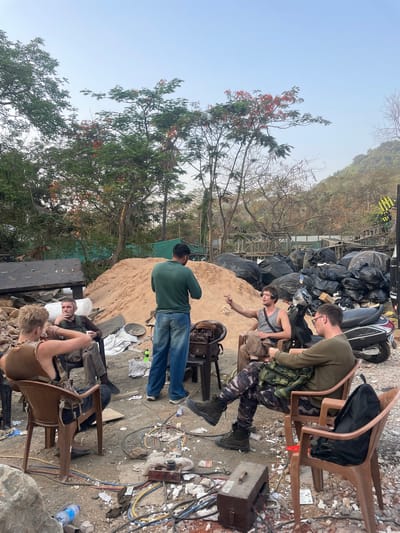 The image depicts an outdoor scene in what appears to be a cluttered or semi-constructed area, possibly within Mira-Bhayander, India, though the location is not explicitly evident.
There are six individuals present. Two are seated in plastic chairs, with one person appearing to adjust or examine something within a bag. Another person stands beside a sand pile, their back to the camera, wearing a green shirt and jeans. A fourth person is sitting in a plastic chair as well. A pile of large black bags and other debris are in the background. Electrical wires, cables, and equipment are scattered on the ground. The environment contains evidence of construction or renovation with exposed wires.