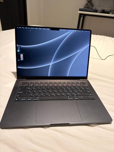 A dark gray MacBook is open and lying on a light-colored, textured surface, likely a bed. The laptop screen displays a bluish-gray abstract wallpaper with flowing curved lines. The keyboard is visible and appears to be in Russian. Several icons are visible in the menu bar at the top of the screen, including a Wi-Fi symbol, battery indicator, and clock showing "20.7. 17:32". To the left of the screen, there are two small thumbnail windows displayed, one above the other. The scene is indoors, with a dark doorway visible in the background on the left. The overall impression is of a personal workspace or leisure setting. The image is captured from a slightly elevated, forward-facing perspective. There is no discernible connection to Ban Phonsavang Neua, Laos.