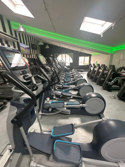 The image shows a row of modern elliptical exercise machines in a gym. The gym features a black and white striped wall in the background, with a bright green LED light strip running along the ceiling. Skylights are visible in the ceiling, allowing natural light to enter the space. Several elliptical machines are lined up, each with a dark gray body and blue accents on the foot pedals and console. The machines appear to be in use, with some showing screens. In the background, other gym equipment and a person can be seen. The scene suggests a well-equipped fitness facility. This gym is located in Enghien-les-Bains, France.