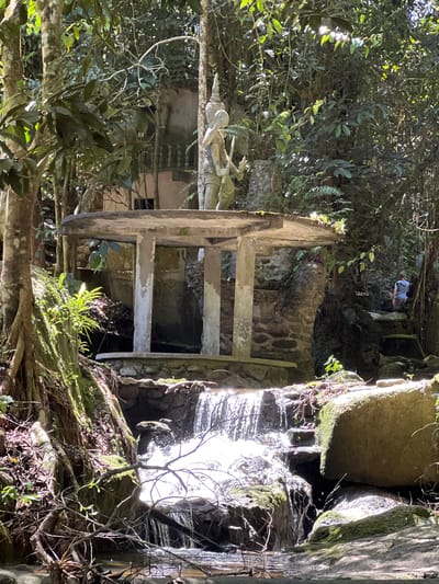 The image depicts a natural scene with a man-made structure in the city of Ban Bang Khao, Thailand. In the foreground, a small waterfall cascades over rocks into a body of water. To the left of the waterfall, large tree roots are visible, covered in moss. Behind the waterfall and roots, a weathered, circular, concrete structure with pillars stands. On top of this structure is a stone statue of a figure holding a weapon. In the background, dense green foliage creates a jungle-like atmosphere. To the right of the concrete structure, a person wearing a blue shirt and grey shorts is walking up a stone staircase. The overall scene is shaded, with sunlight filtering through the trees.