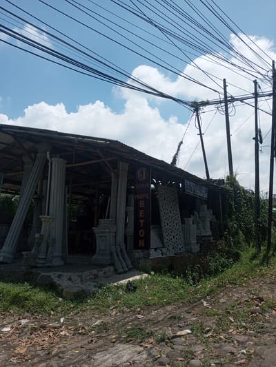 The image depicts a roadside concrete ornament shop in an outdoor setting. The shop, with a simple corrugated roof supported by wooden beams, displays various concrete pillars, panels, and bases. A sign on the side reads "SAKINAH PROFIL BETON." Power lines crisscross above the shop and adjacent utility poles. Vegetation grows around the base of the shop and along the roadside. The ground is a mix of dirt, rocks, and scattered leaves. The sky is partly cloudy. The location is not identifiable as Kioumaknao 2, Laos.